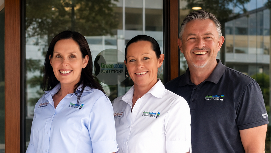 Three employees standing together in front of a glass office entrance. Two women wear matching white and navy blue uniforms, and one man wears a navy polo shirt with a logo and khaki pants.